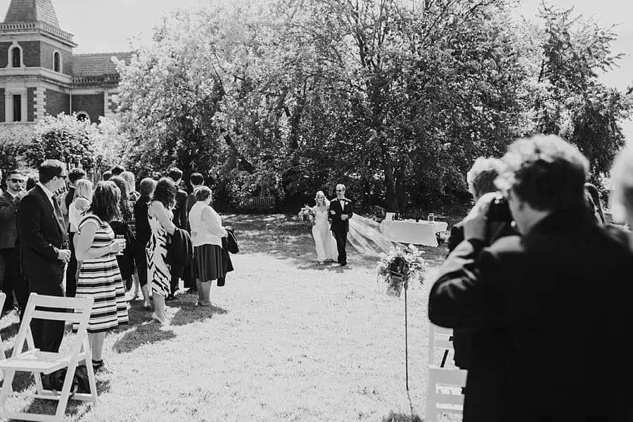 Black-and-white wide view of guests watching the bride approach across a lawn.