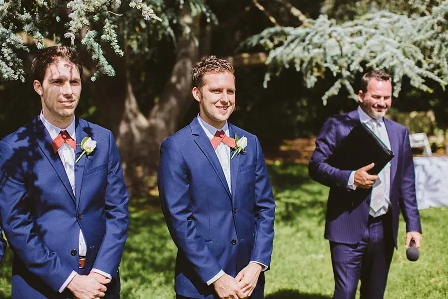 Groom and a suited companion waiting beneath a large tree.