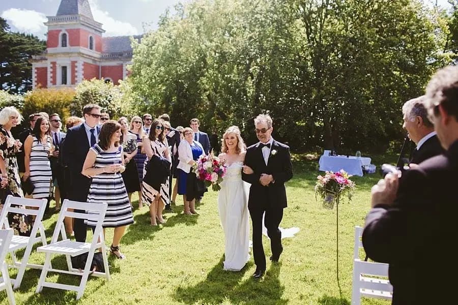 Bride walking beside a suited man through rows of standing wedding guests.