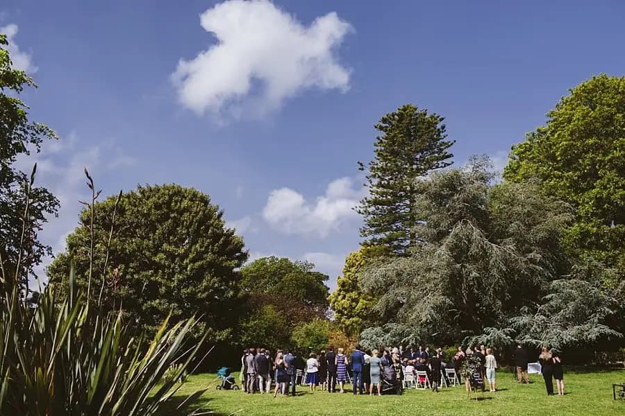 Wide view of the garden ceremony beneath trees and a blue sky.