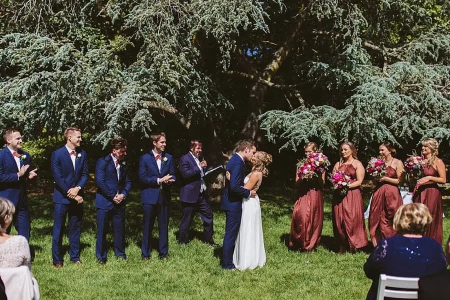 Wedding party applauding as the bride and groom kiss beneath a tree.