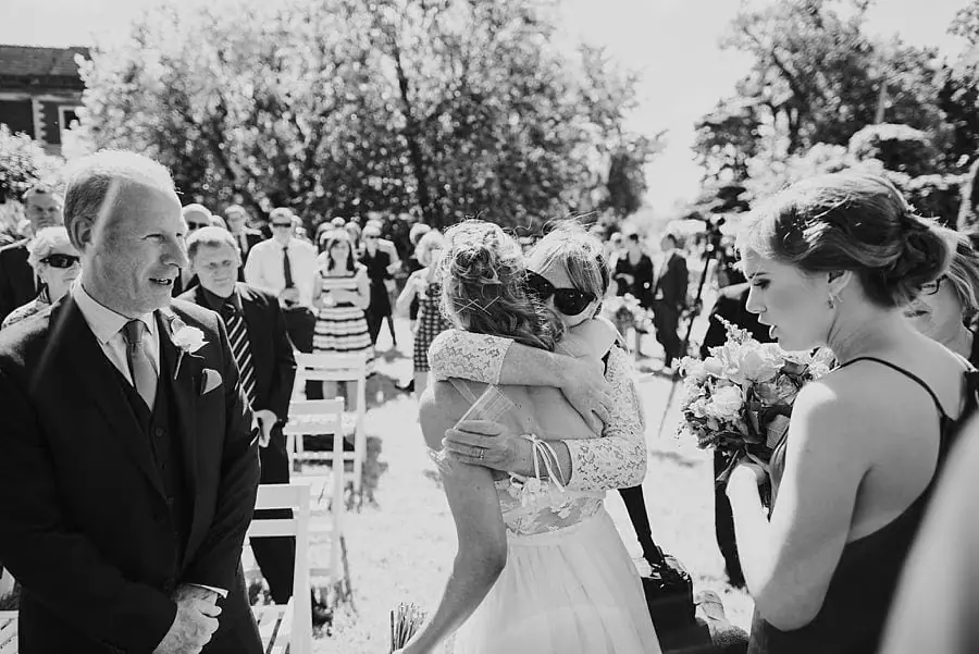 Black-and-white photograph of the bride embracing a woman beside ceremony chairs.
