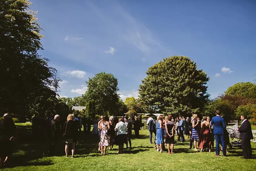 Wedding guests mingling on a sunny lawn beneath large trees.