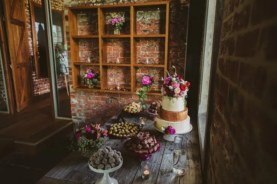 Wedding cake and trays of sweets displayed beside a brick wall and wooden shelving.