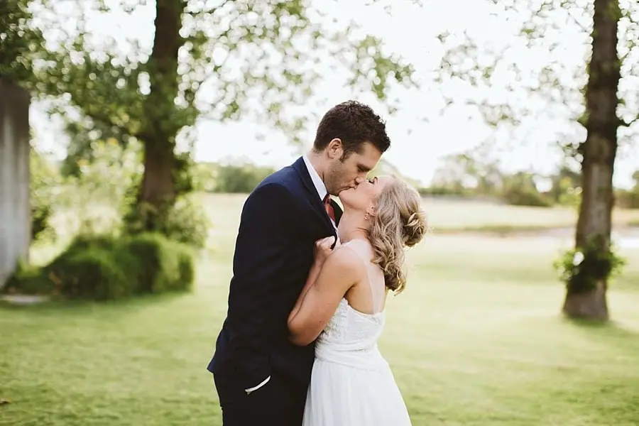 Groom kissing the bride as they stand on a lawn.
