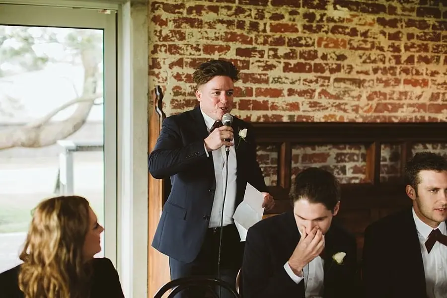 Suited man speaking into a microphone beside seated reception guests.