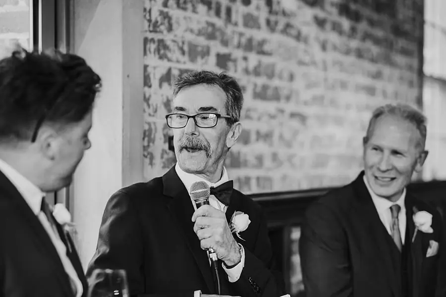Black-and-white photograph of a man with glasses speaking into a microphone.