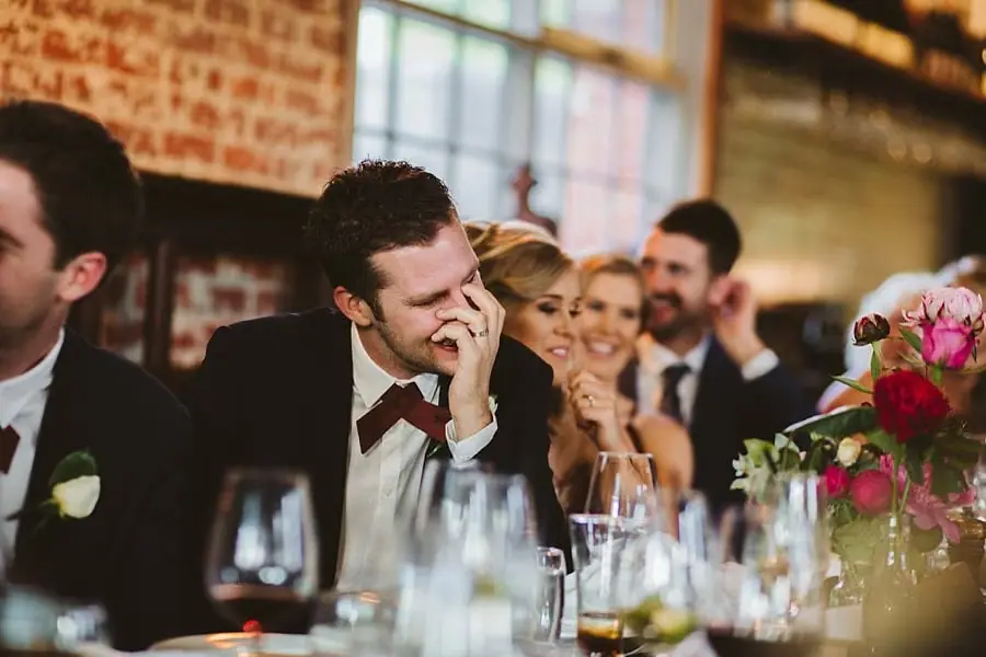 Groom smiling with a hand near his face as the bride sits beside him.