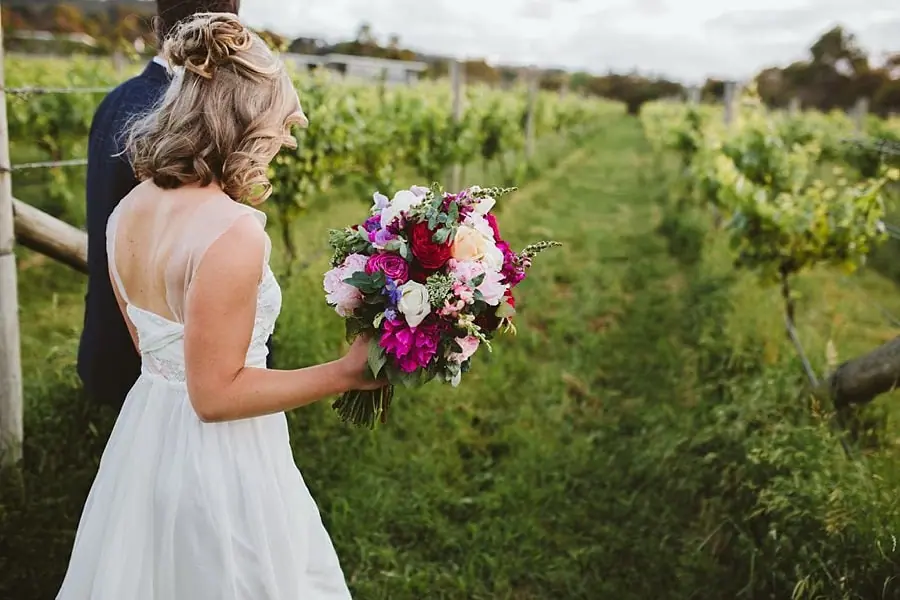 Bride holding her bright bouquet while walking among vineyard rows.