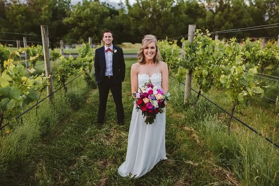 Bride and groom standing apart along a narrow path between vines.