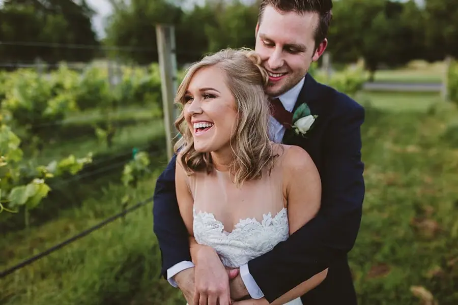 Bride laughing as the groom embraces her from behind among vines.