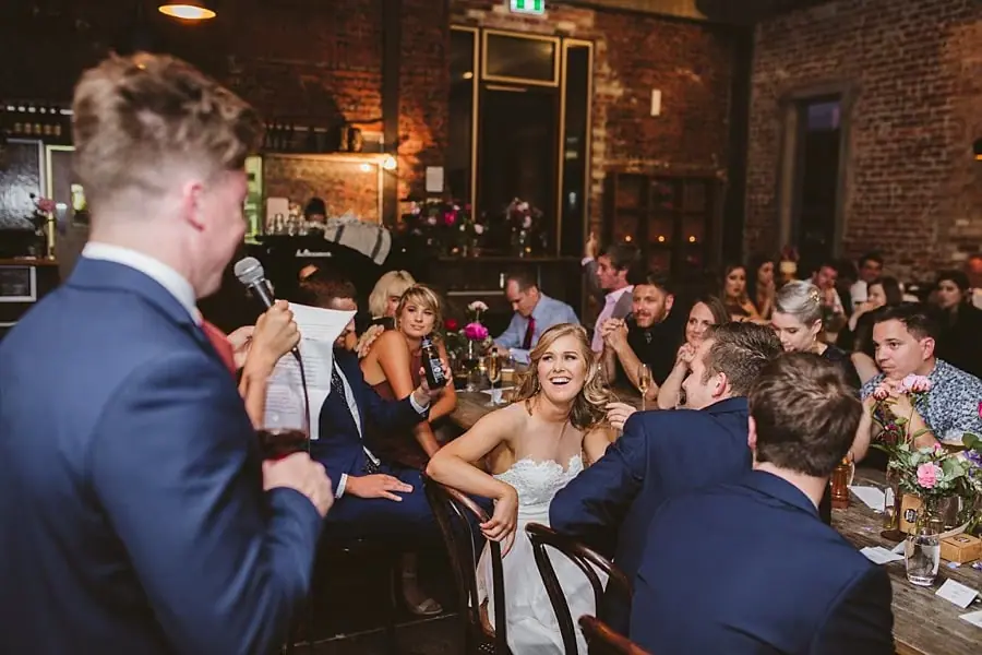 Suited man reading a speech as the couple and guests laugh at their table.