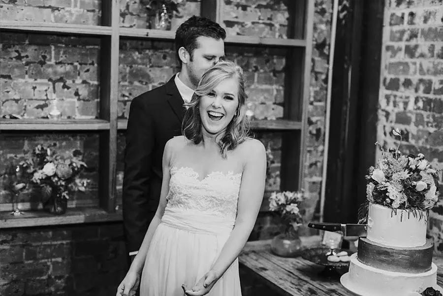 Black-and-white photograph of the groom kissing the bride's head beside their cake.
