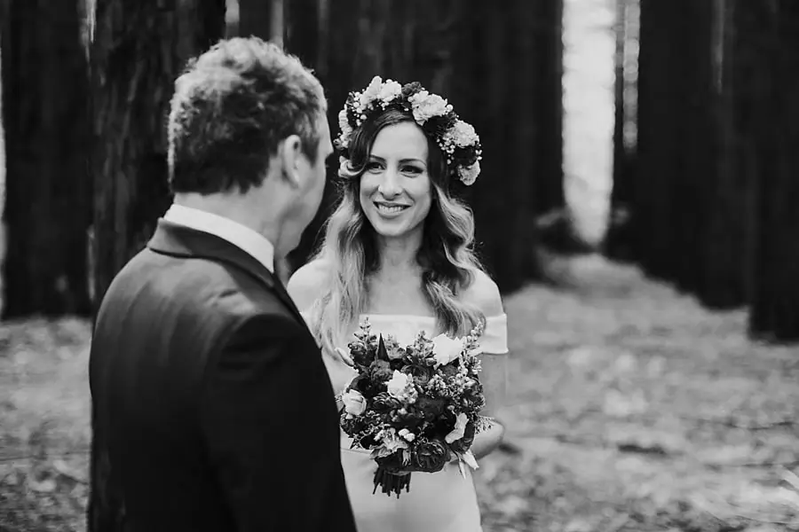 Black-and-white photograph of a bride smiling at her groom while holding a bouquet among redwood trees.
