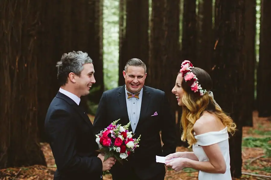 Couple holding hands beside a celebrant among tall forest trees.