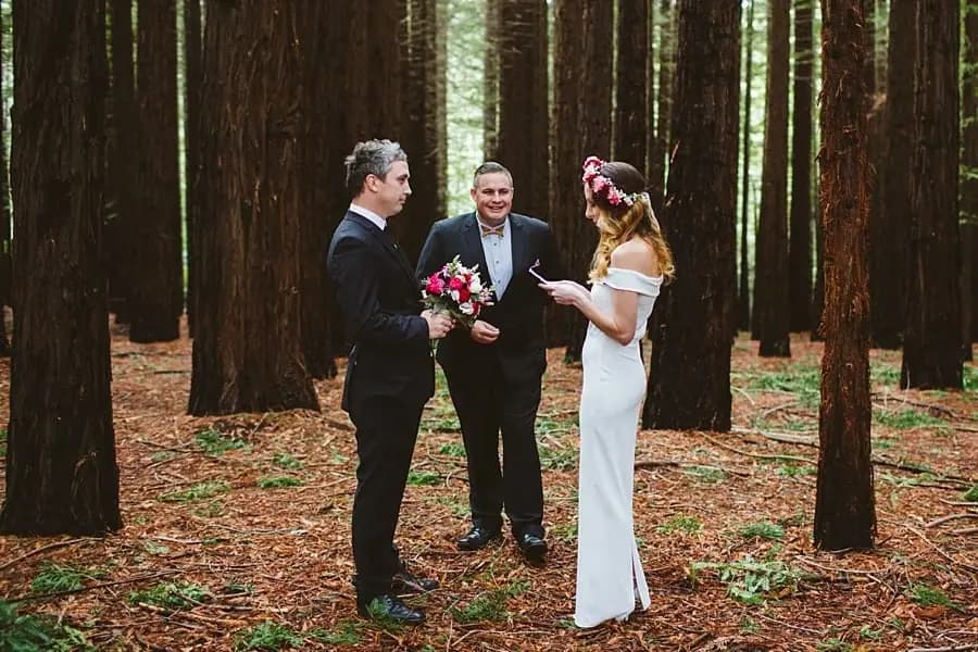 Wide view of a bride, groom and celebrant during a ceremony among redwood trees.
