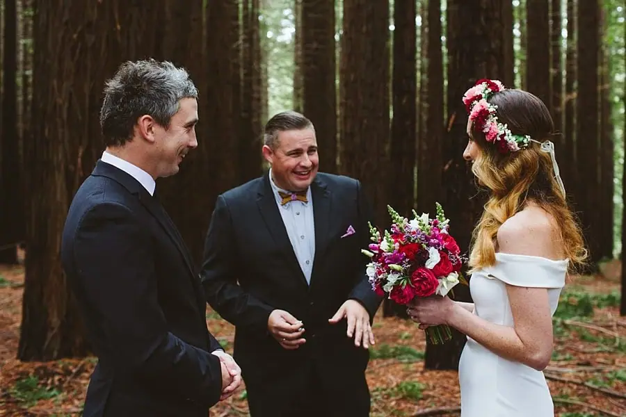 Bride and groom standing beside their celebrant with a red and white bouquet between them.