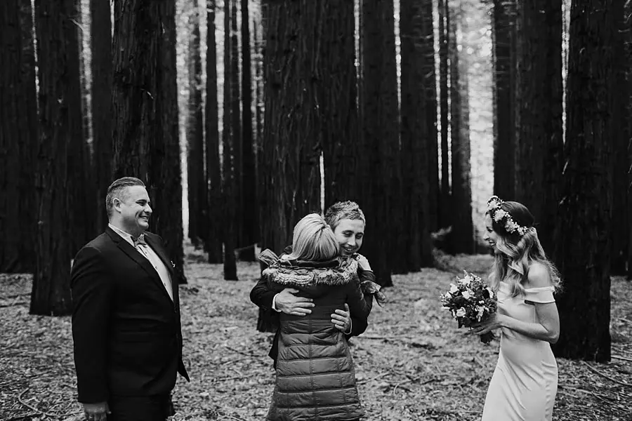 Black-and-white photograph of the celebrant hugging a guest as the newlyweds look on beneath redwood trees.
