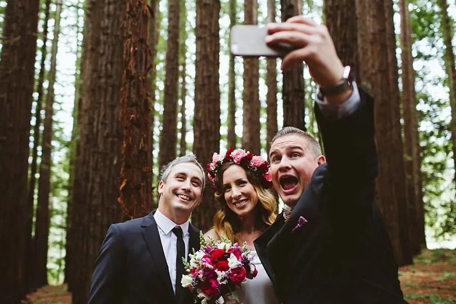 Celebrant holding up a phone for a selfie with the smiling newlyweds in the forest.