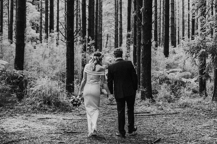 Black-and-white photograph of a couple walking hand in hand away through the forest.