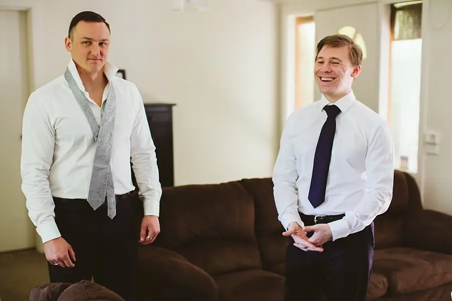 Two men in white shirts and ties smiling indoors before the wedding.