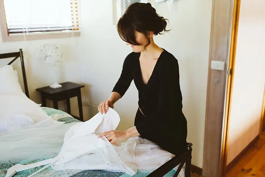 Bride in a black robe lifting her white wedding dress from a bed.