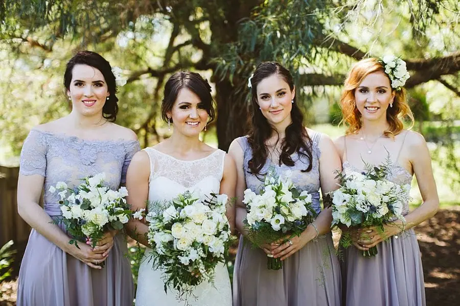 Bride and three women in pale dresses holding white bouquets beneath a tree.