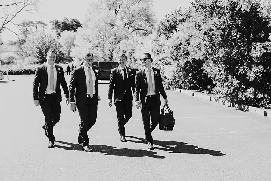 Black-and-white photograph of four suited men walking across a sunlit car park.