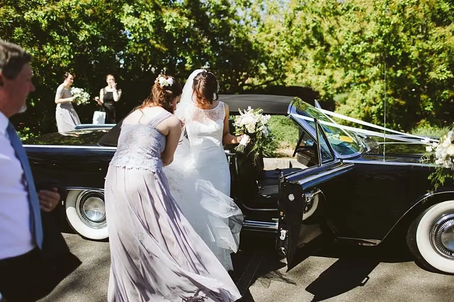 Bride stepping from a black convertible with help from a woman in a pale dress.