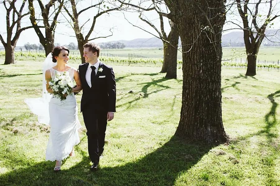 Bride and groom walking together through sunlit trees.