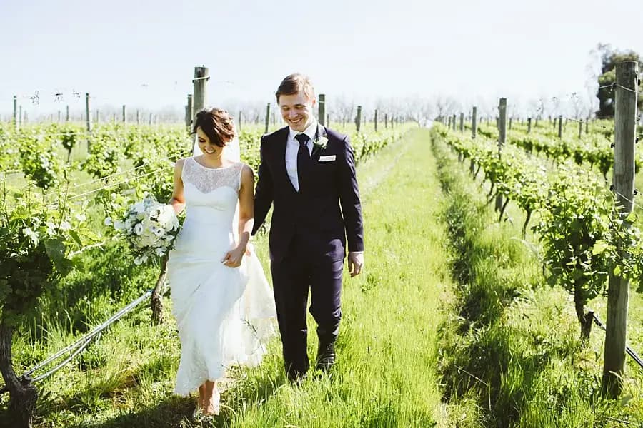 Bride and groom walking hand in hand through a sunlit vineyard.
