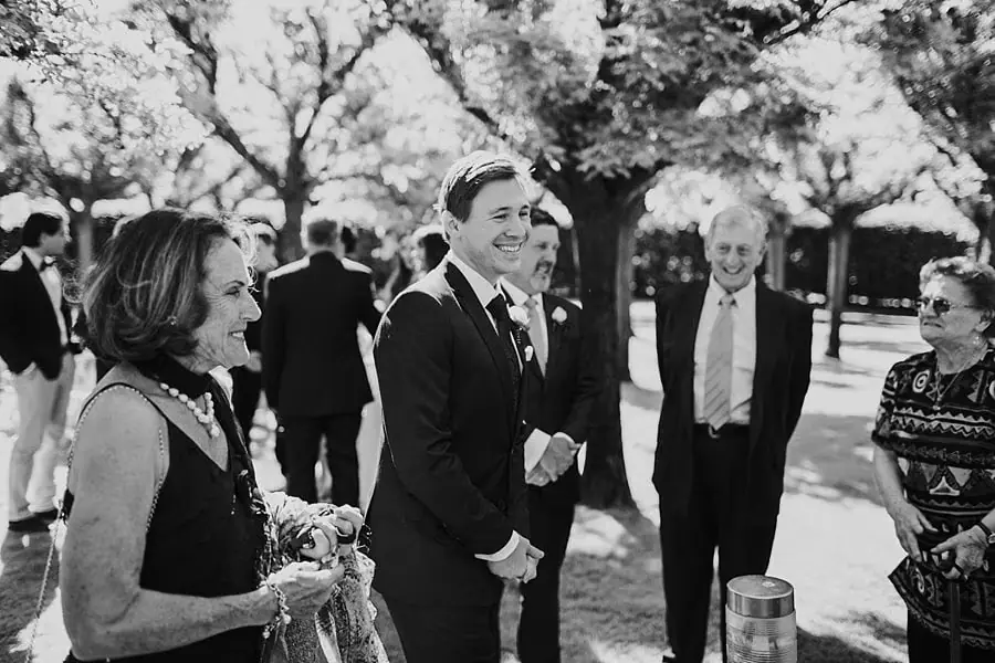 Black-and-white photograph of the groom standing with guests beneath trees.