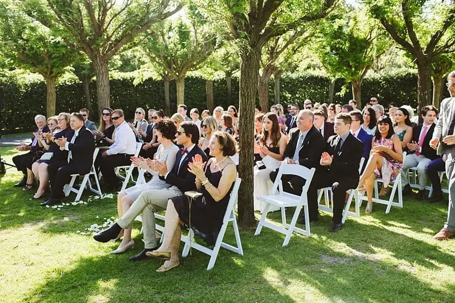 Guests sitting in white chairs at an outdoor ceremony beneath leafy trees.