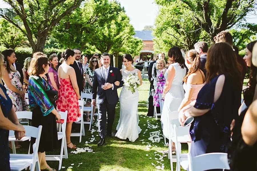 Bride walking along the outdoor ceremony aisle with a suited man as guests stand beside them.