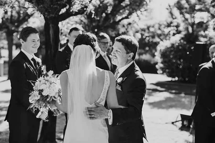 Black-and-white photograph of a suited man beside the bride as she faces the groom.