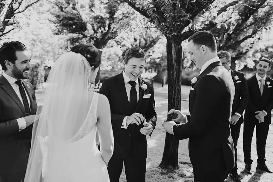 Black-and-white photograph of men exchanging a small item beside the bride during the ceremony.