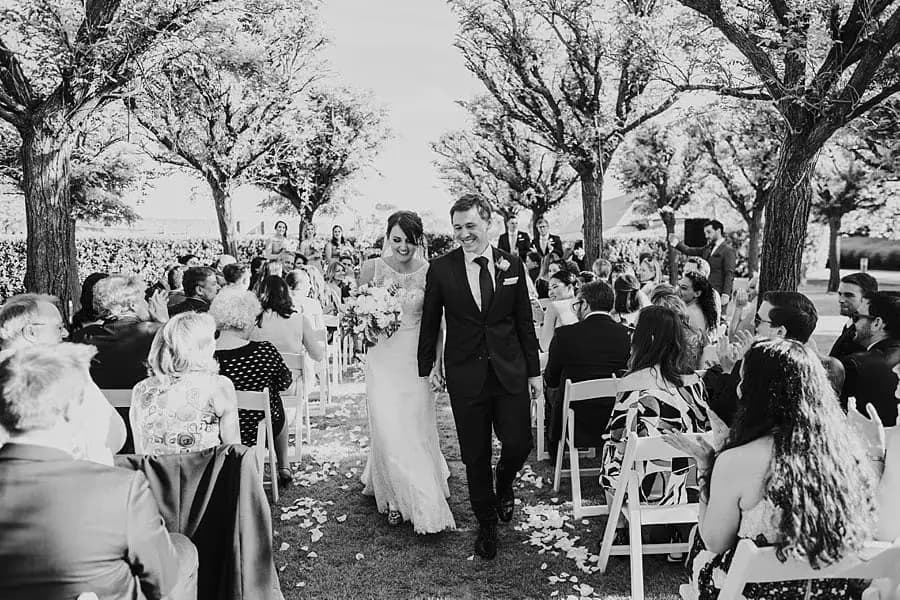 Black-and-white photograph of the newlyweds walking down the aisle as guests applaud.