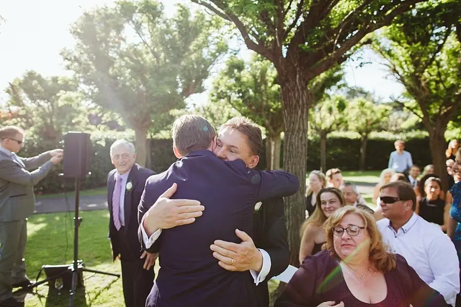 Groom embracing a guest beneath a sunlit tree.