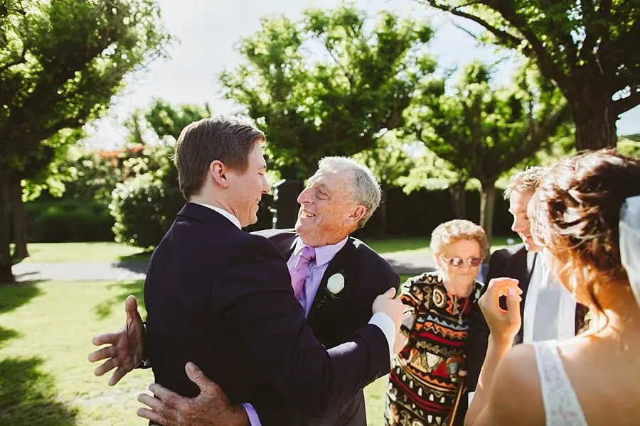 Groom greeting an older suited guest beside the bride outdoors.