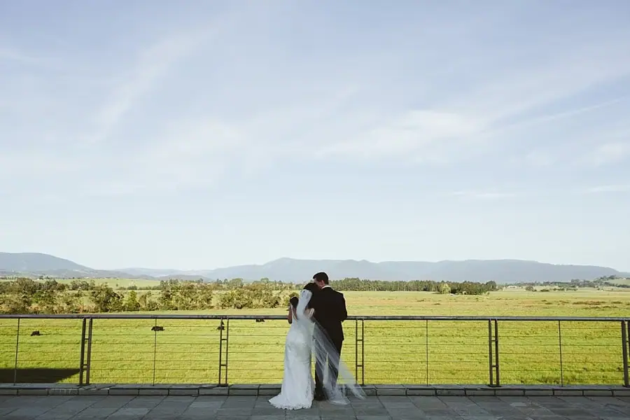 Bride and groom embracing at a railing overlooking open fields and distant hills.
