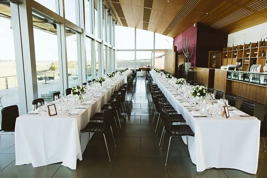 Two long reception tables set with white cloths, flowers and glasses in a window-lined room.