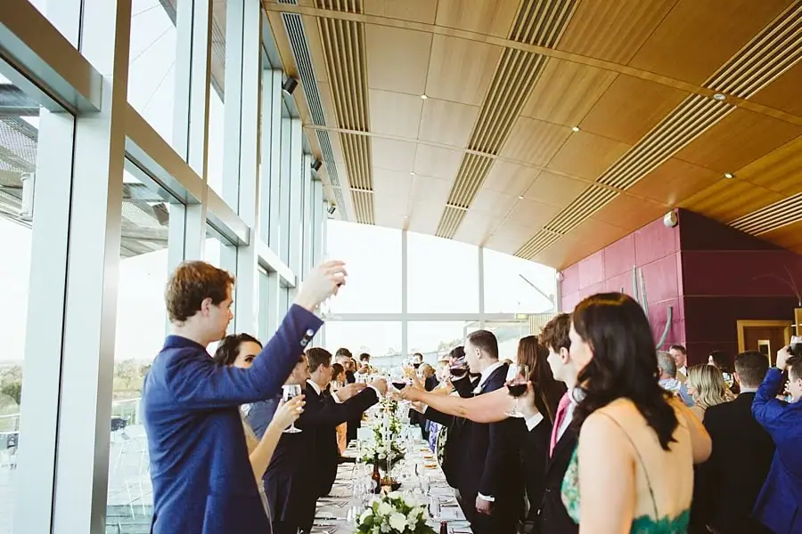 Guests raising their glasses around a long reception table beside tall windows.