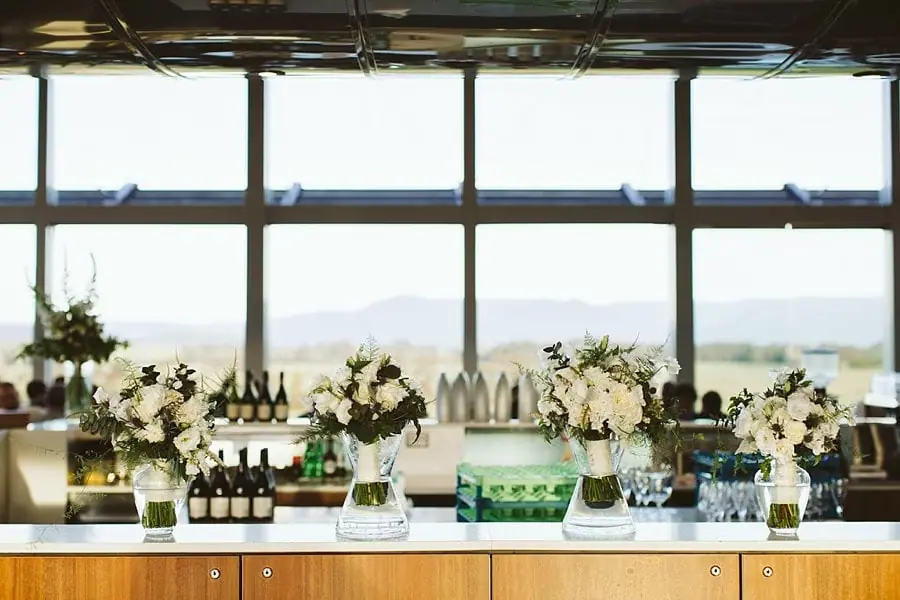 White flower arrangements decorating a reception bar beneath large windows.