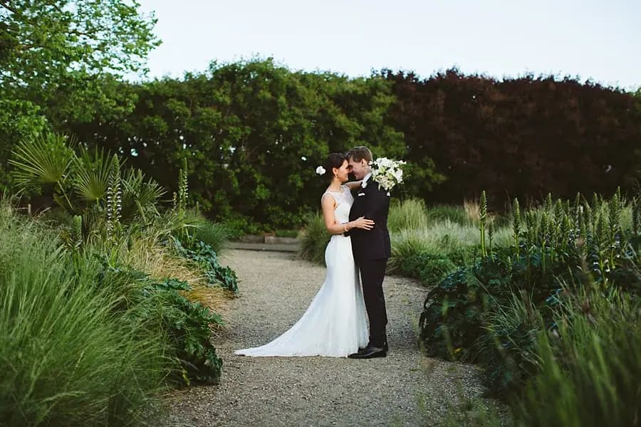 Bride and groom kissing on a gravel path surrounded by garden plants.