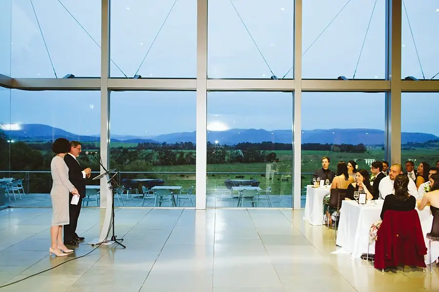 Woman speaking into a microphone beside the groom as reception guests watch.