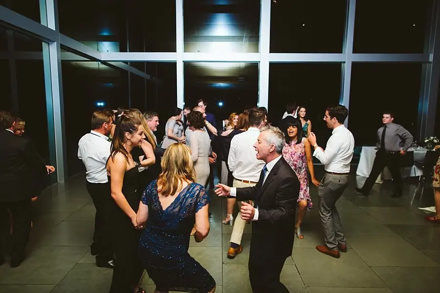 Wedding guests dancing together beside tall reception windows.