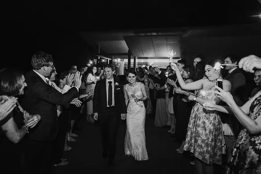 Black-and-white photograph of the newlyweds walking between applauding reception guests.