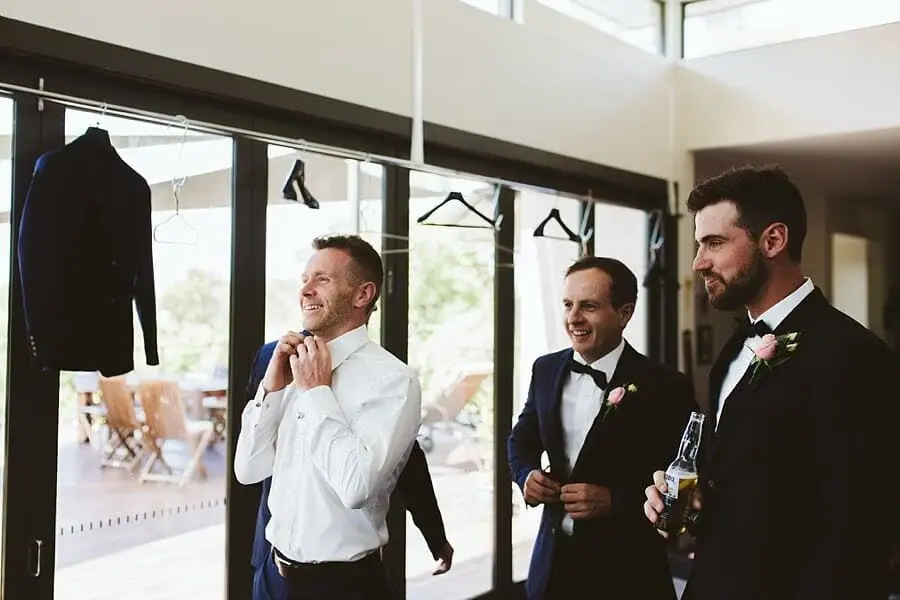 Groom adjusting his black bow tie while two suited men smile beside him.