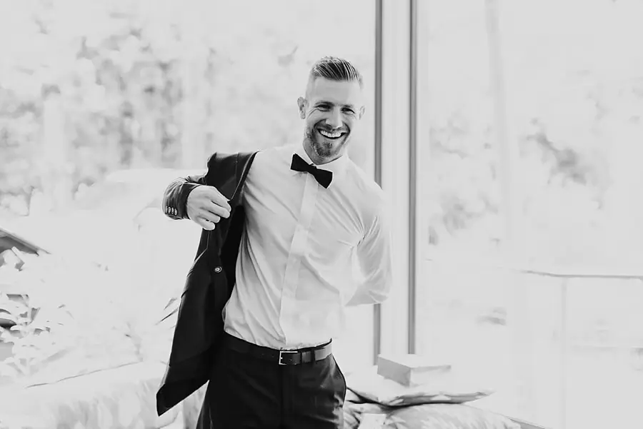 Black-and-white photograph of the smiling groom putting on his suit jacket.