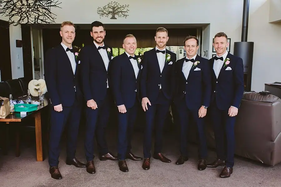 Six men in dark suits and bow ties posing together indoors.